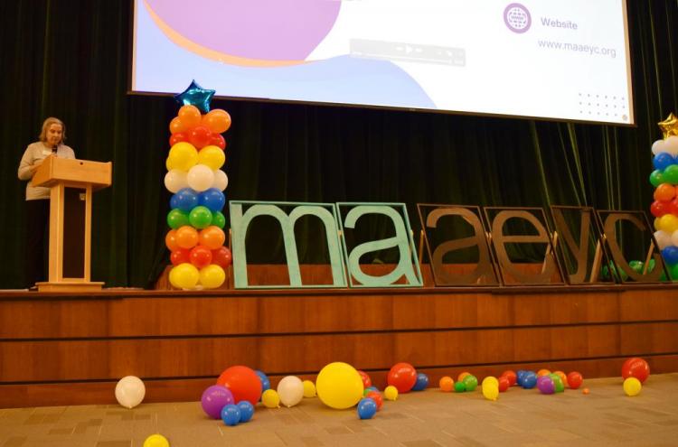 woman stands at podium speaking, next to giant MAAEYC sign